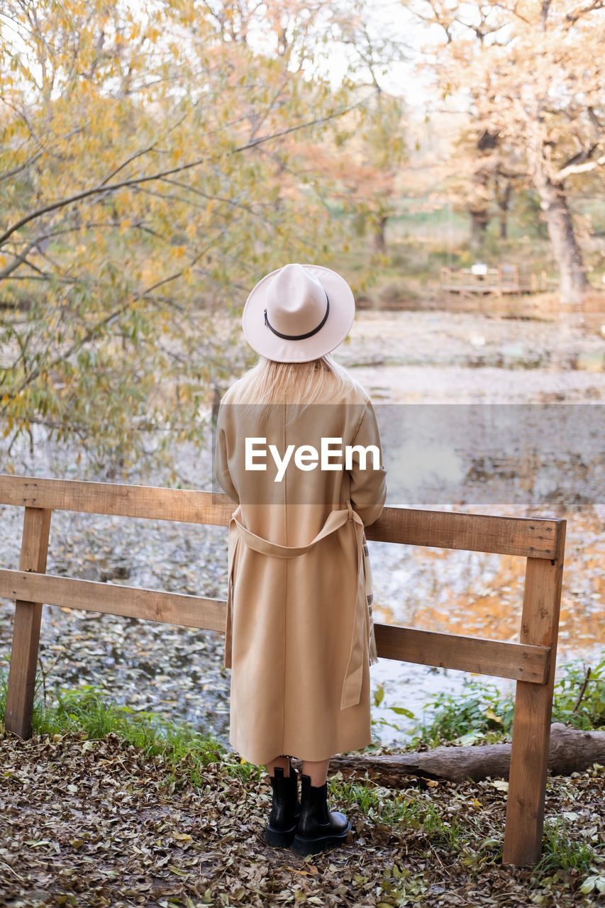 Rear view of beautiful woman in beige coat and hat by the lake in autumn park in fall