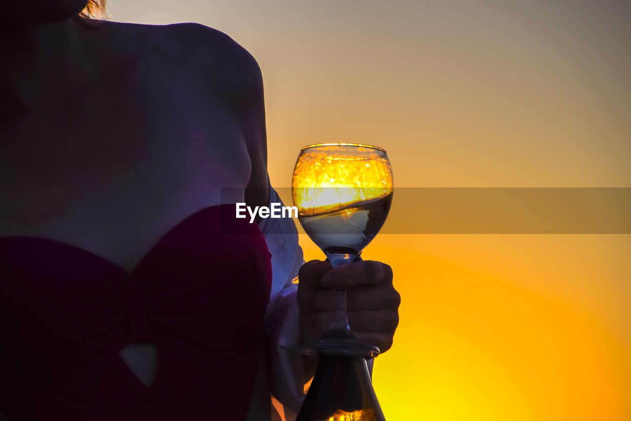 CLOSE-UP OF MAN HOLDING GLASS AGAINST SUNSET SKY