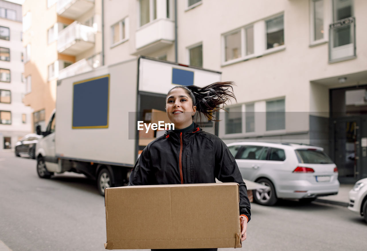 Confident young delivery woman carrying cardboard box on street in city