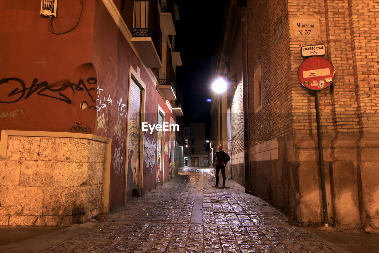 REAR VIEW OF WOMAN WALKING ON FOOTPATH AMIDST ILLUMINATED BUILDINGS