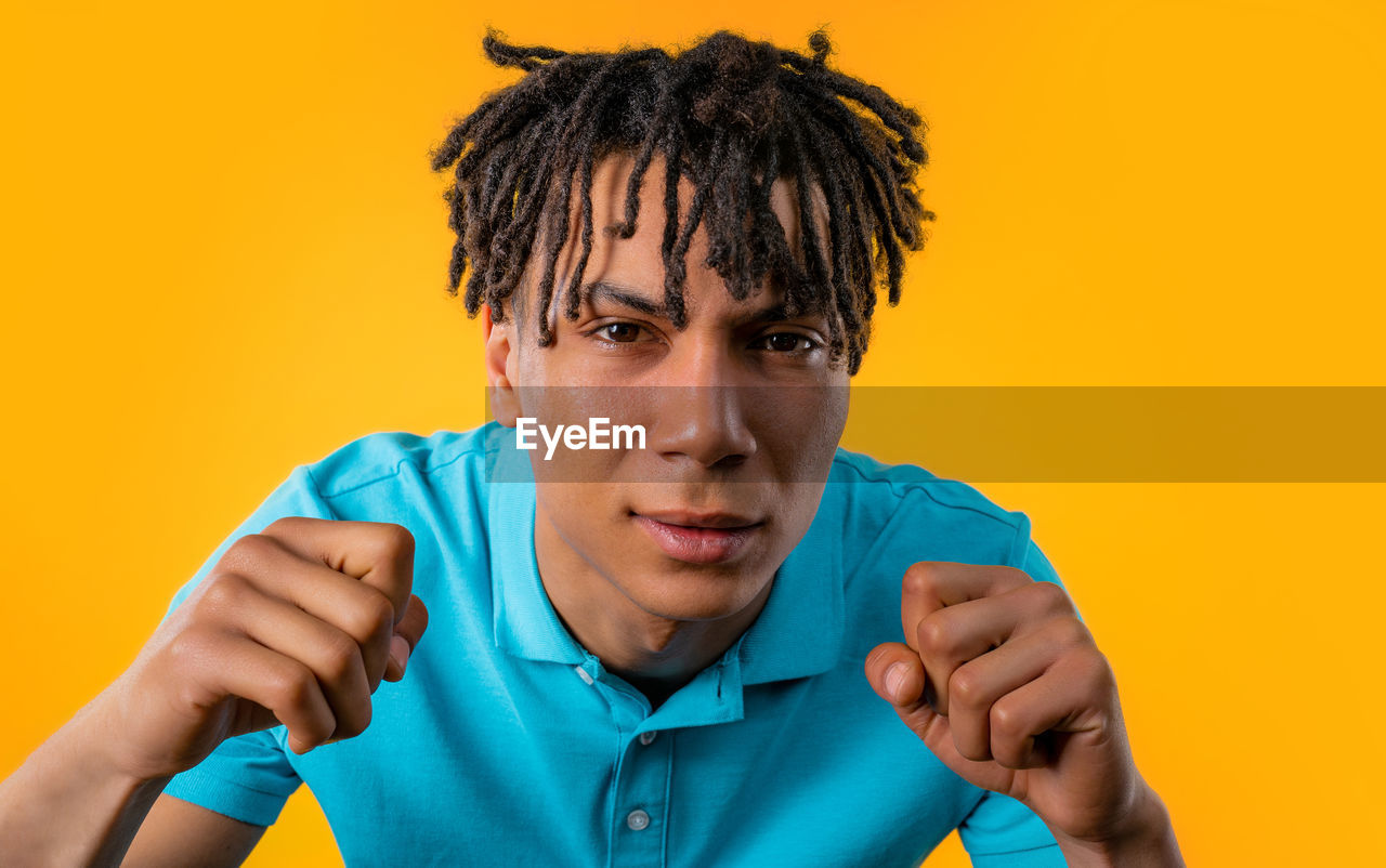 colored background, studio shot, portrait, one person, yellow, yellow background, adult, men, person, emotion, teenager, headshot, indoors, young adult, front view, looking at camera, orange color, human face, facial expression, orange background, negative emotion, casual clothing, lifestyles, clothing, blue, hand, frustration, anger, copy space, hairstyle