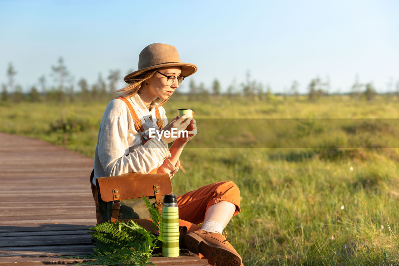 Naturalist woman resting on boardwalk, drinking tea, enjoying the moment at sunset. ecotourism