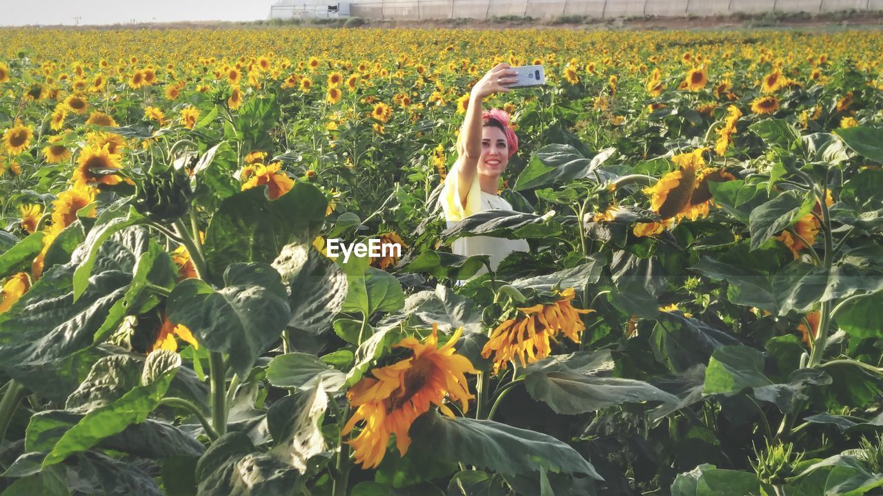 Smiling woman taking selfie through mobile phone while standing amidst flowering plants on field