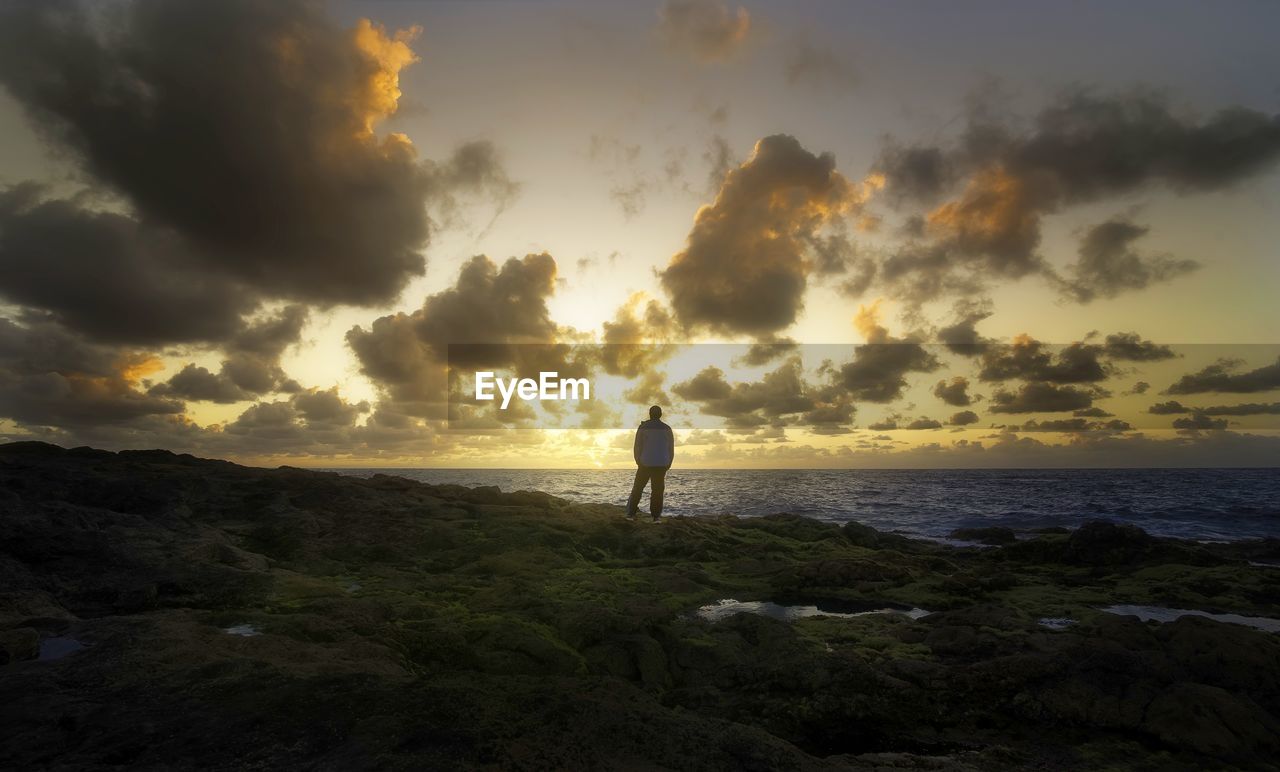 MAN STANDING ON SHORE AGAINST SEA DURING SUNSET