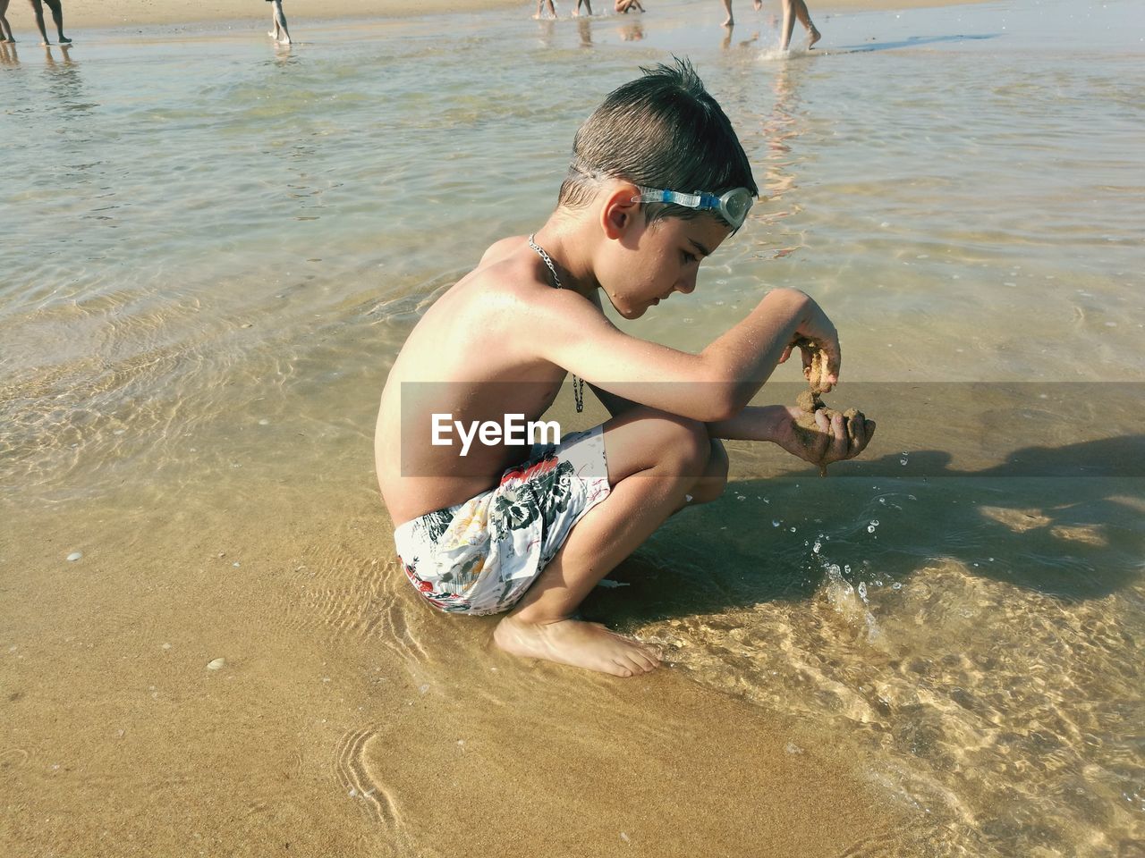 Shirtless boy playing with sand while crouching at beach