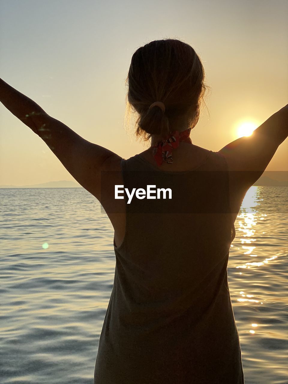 REAR VIEW OF WOMAN STANDING AGAINST SEA AT SUNSET