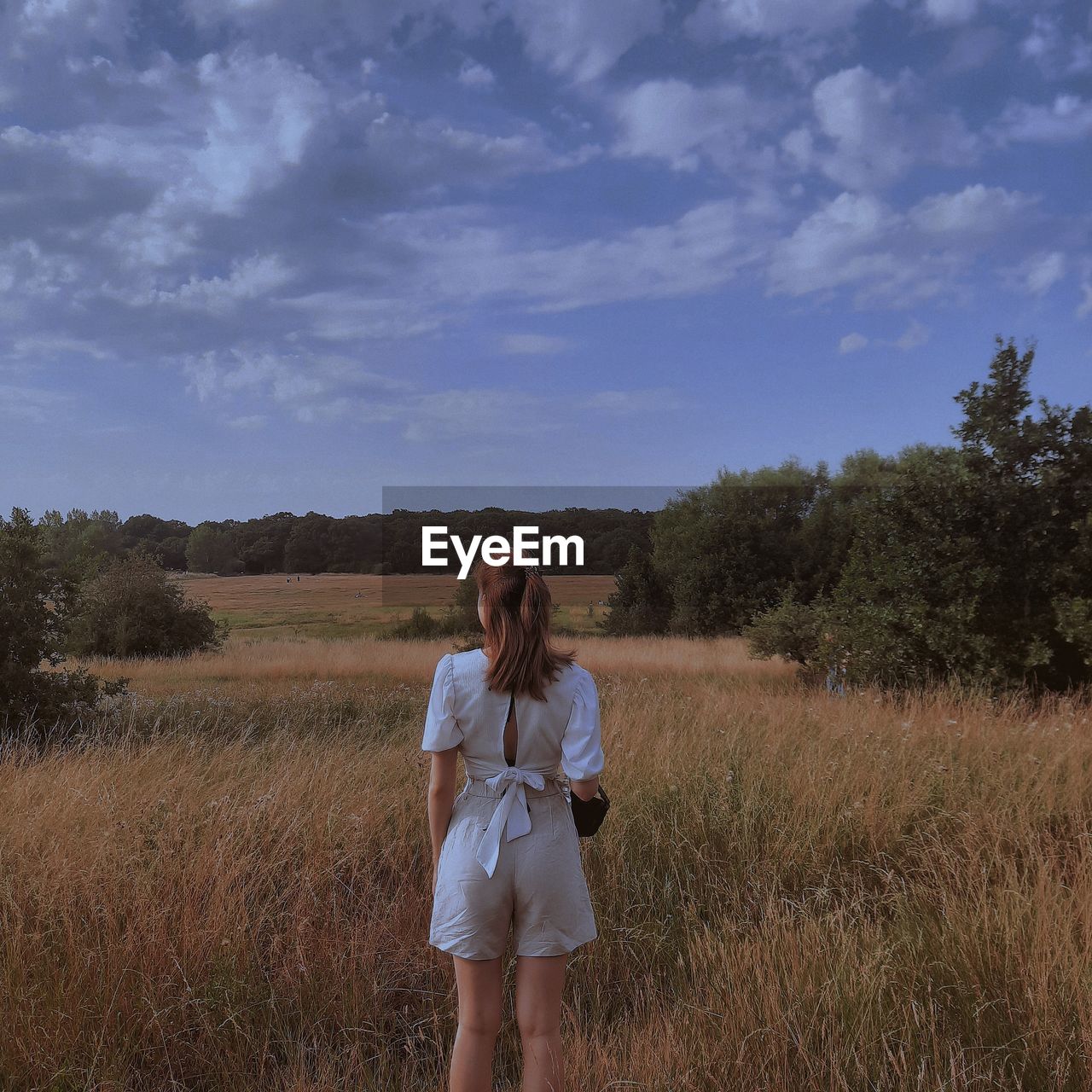 WOMAN STANDING ON FIELD AGAINST TREES AND SKY