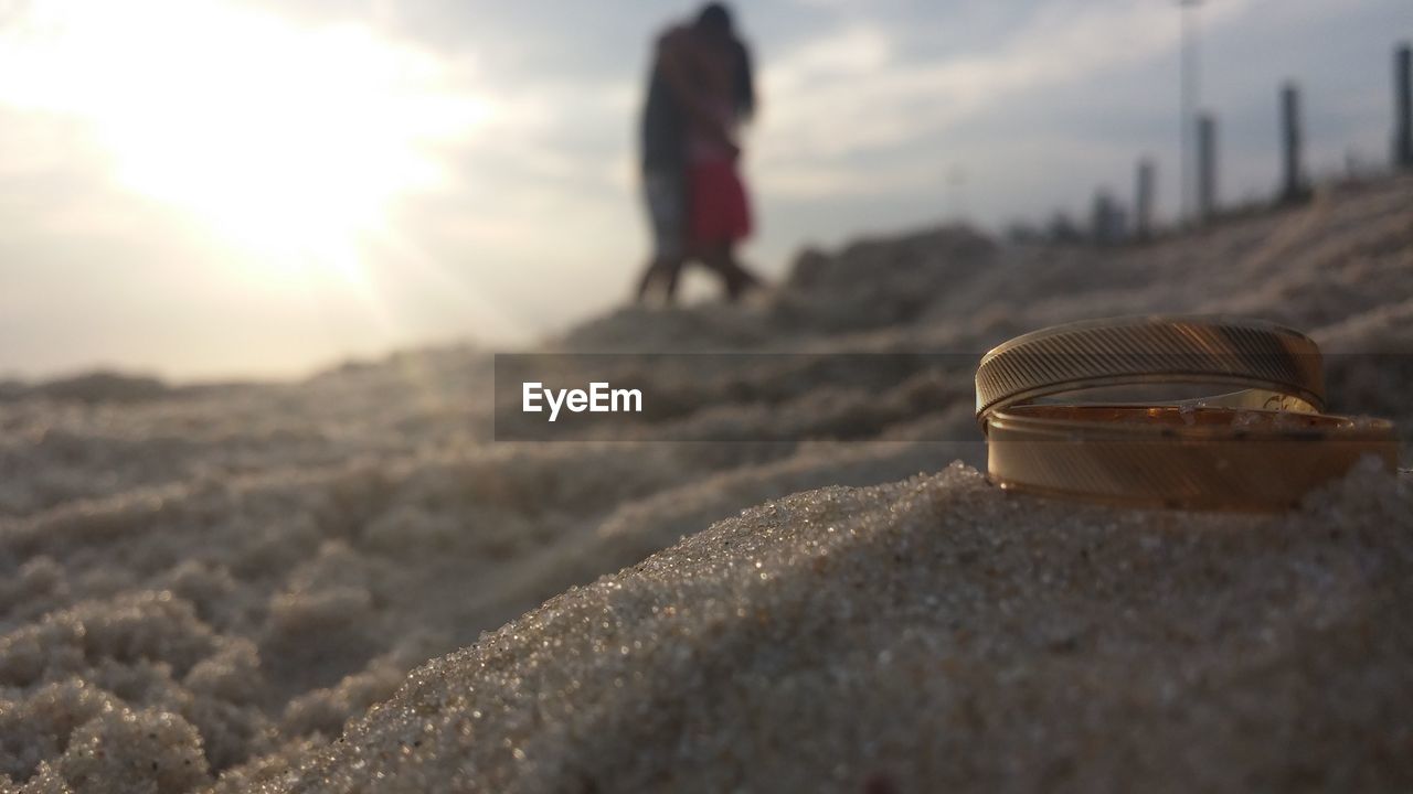 Close-up of wedding rings on sand with couple in background