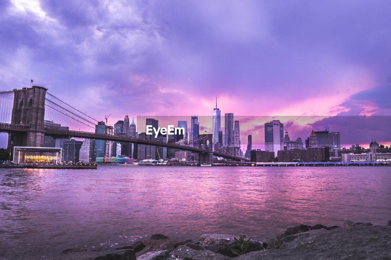 Bridge over river by buildings against sky at dusk