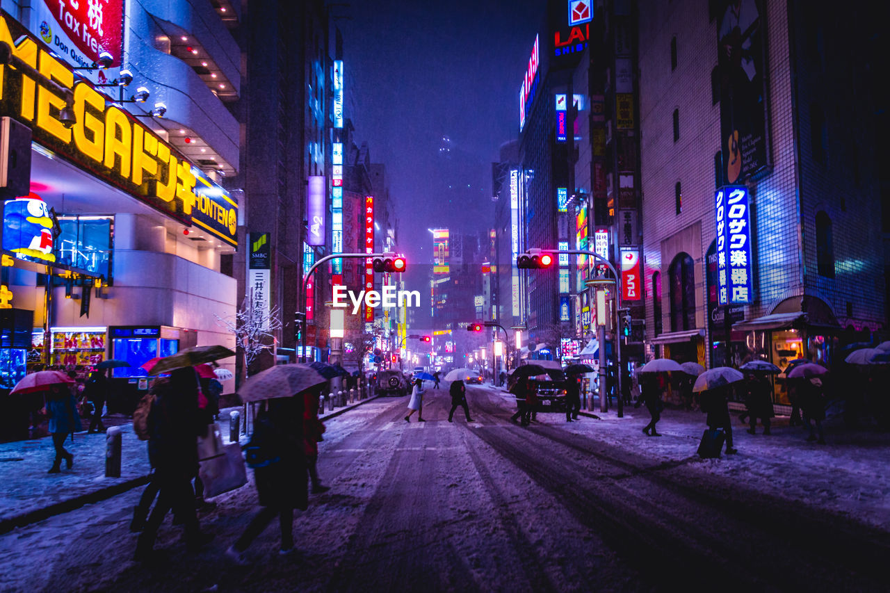 People walking on illuminated street amidst buildings during snowfall