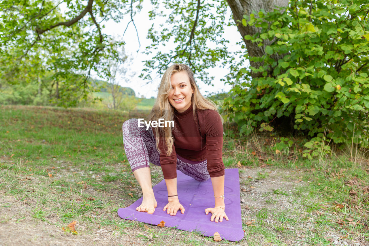 Smiling young woman sitting in a yoga pose in a park on a yoga mat with a tree on a background