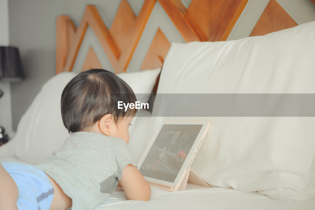 REAR VIEW OF BOY USING MOBILE PHONE ON BED