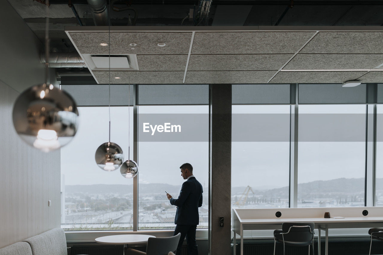 Rear view of businessman with smart phone standing by window at office