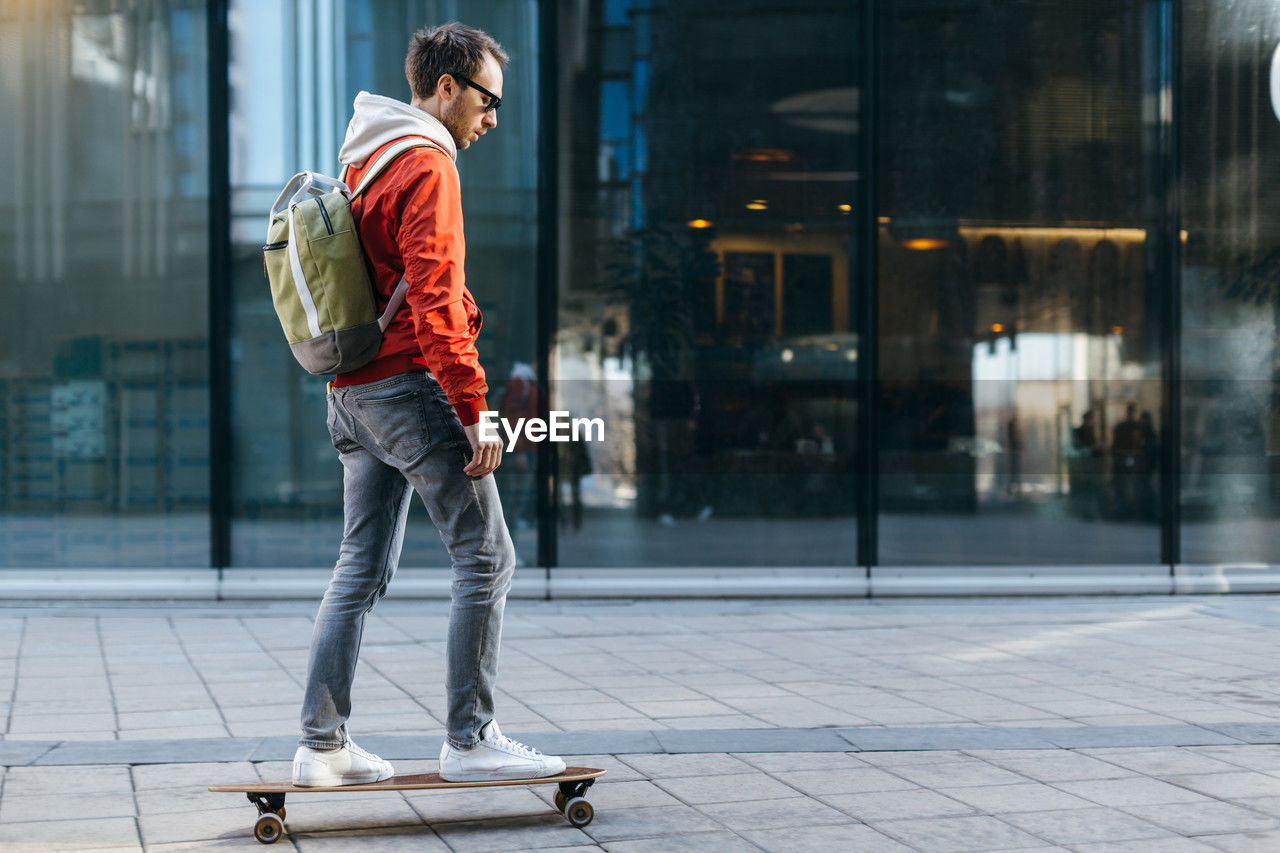 Stylish hipster in red jacket, sunglasses, jeans riding on longboard. concept of leisure activity