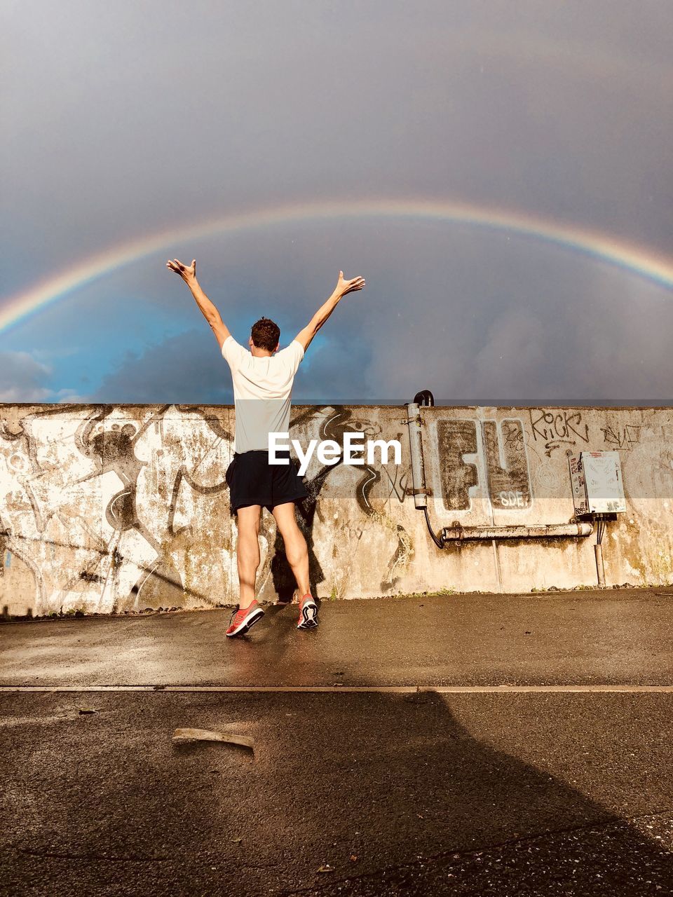 Rear view of man with arms raised standing on road against graffiti wall