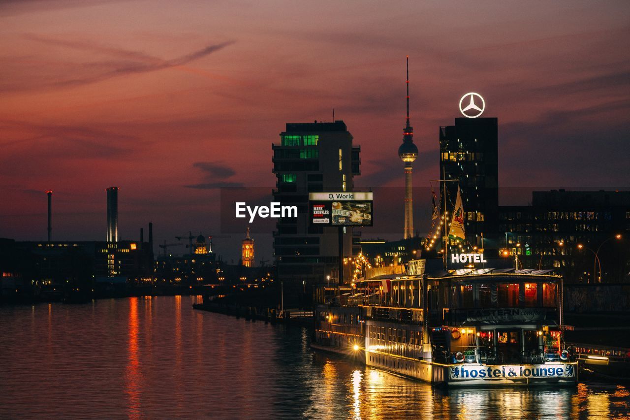 Illuminated fernsehturm and skyscrapers by river against sky during sunset