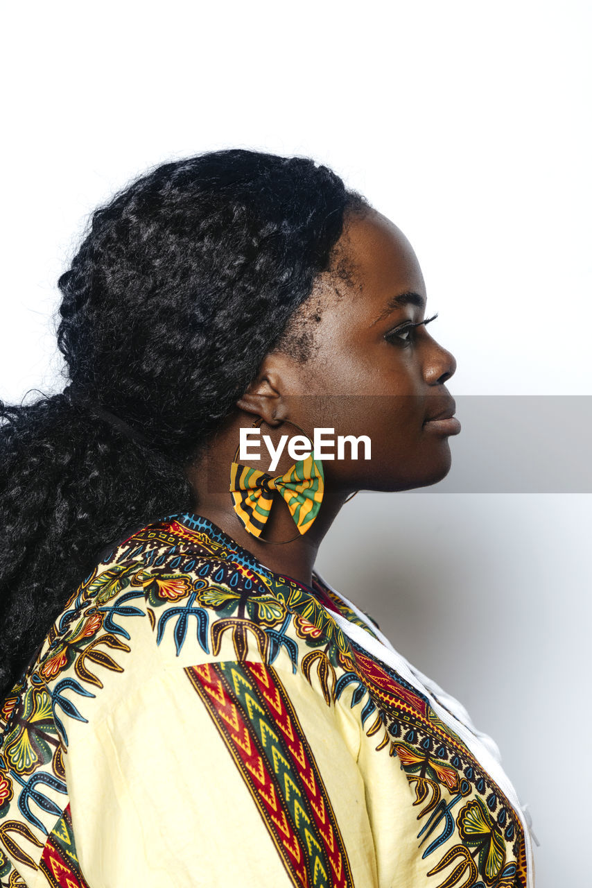 Side view of young curvy woman with afro hairstyle wearing colorful african shirt and earrings. she is looking away while standing on white wall background