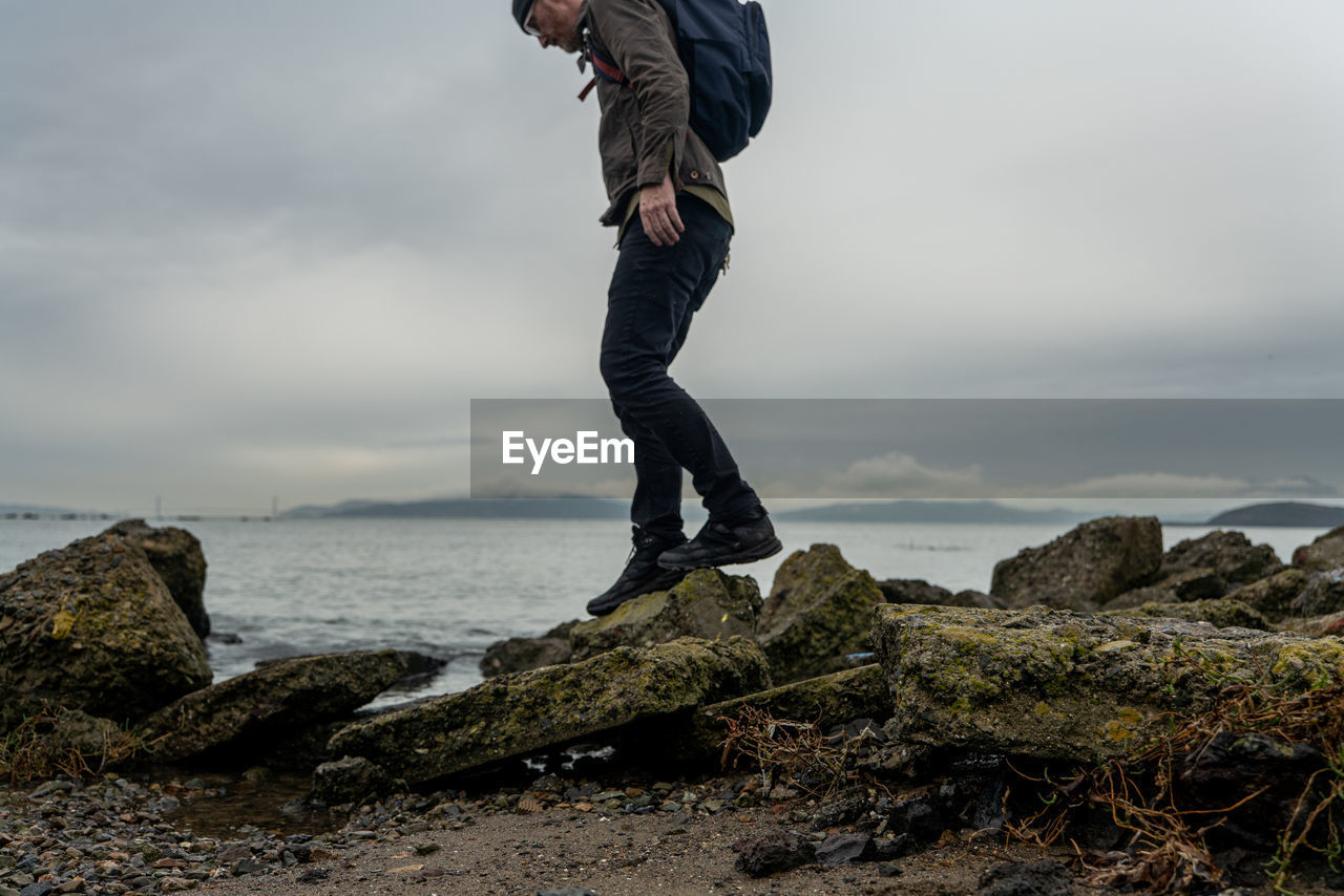 Man with backpack climbs small path of rocks beside bay under gray sky