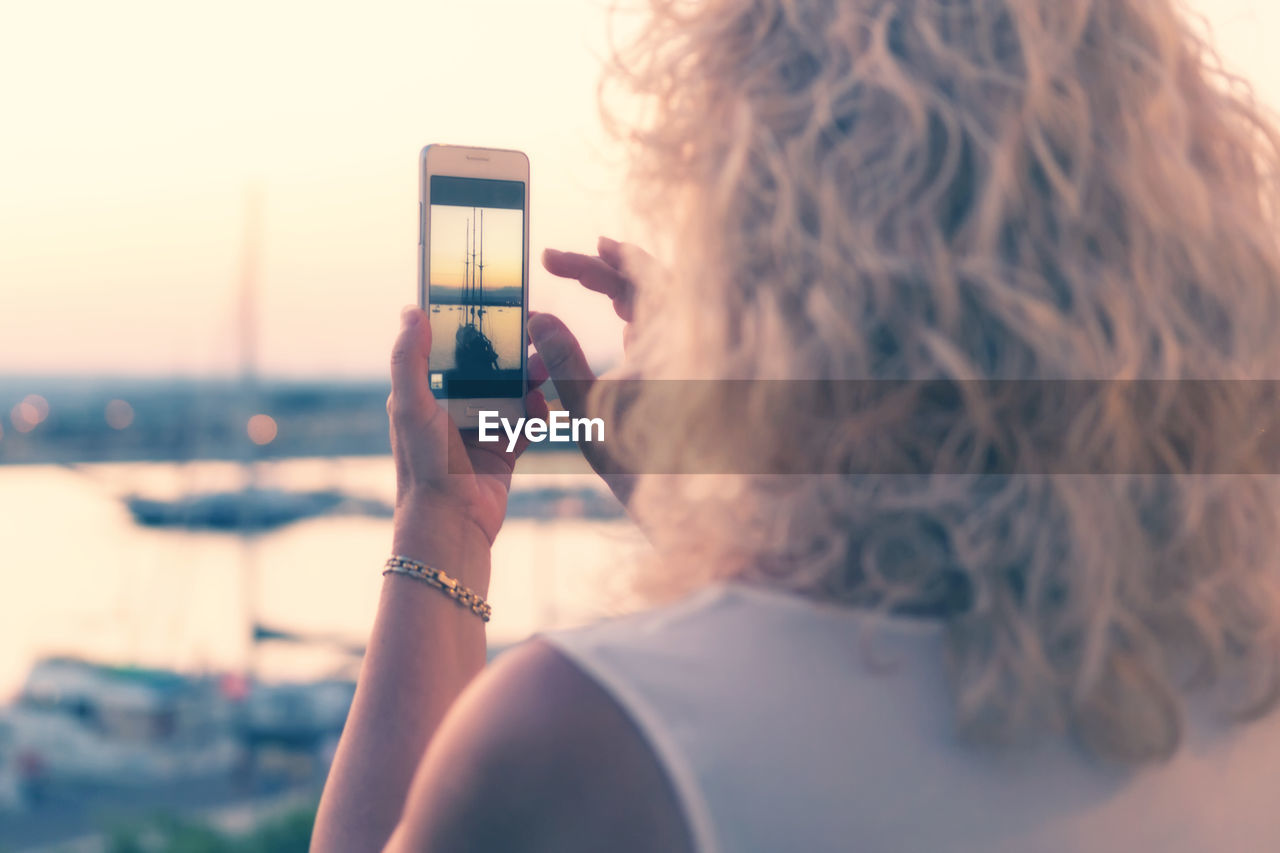 REAR VIEW OF WOMAN PHOTOGRAPHING WITH MOBILE PHONE IN BACKGROUND