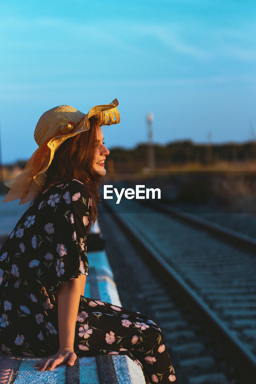 Side view of smiling young woman wearing hat sitting on railroad station platform against sky during sunset