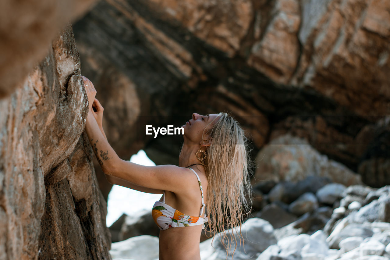 Side view of woman wearing bikini with eyes closed standing by rock formation