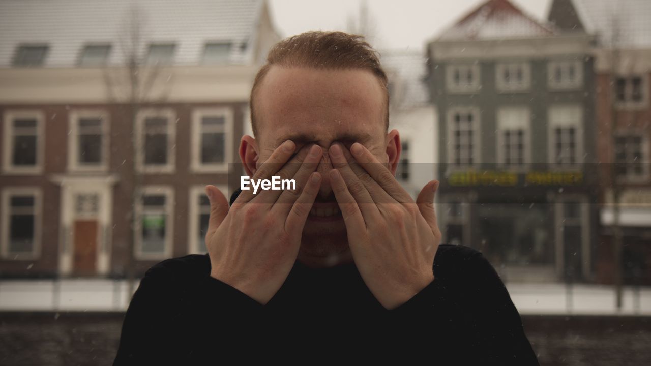 PORTRAIT OF PERSON COVERING FACE WITH HAND AGAINST BUILDINGS