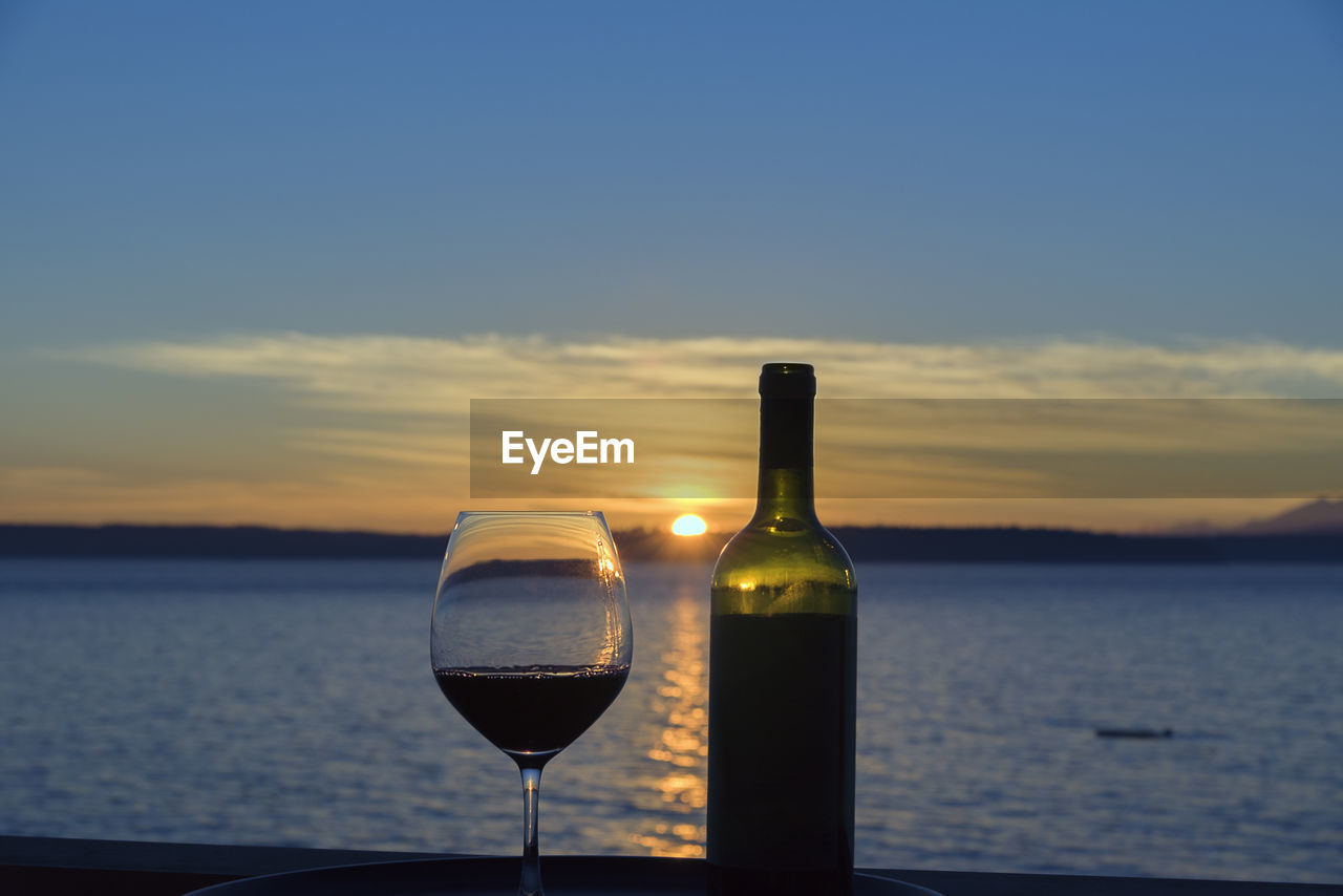 Close-up of wineglass and bottle against sea during sunset