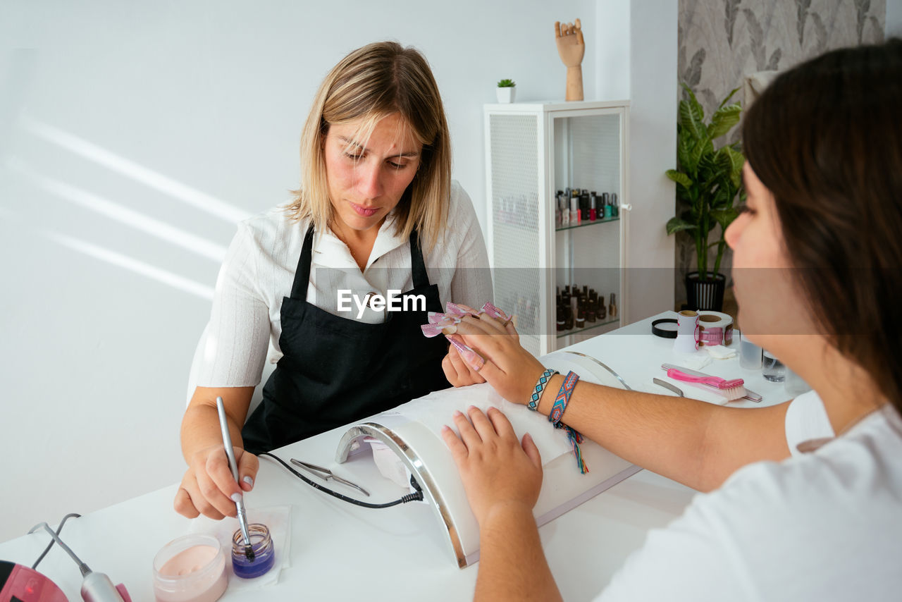 From above of attentive adult female master in apron doing nail extension for anonymous customer sitting at table near window in light salon