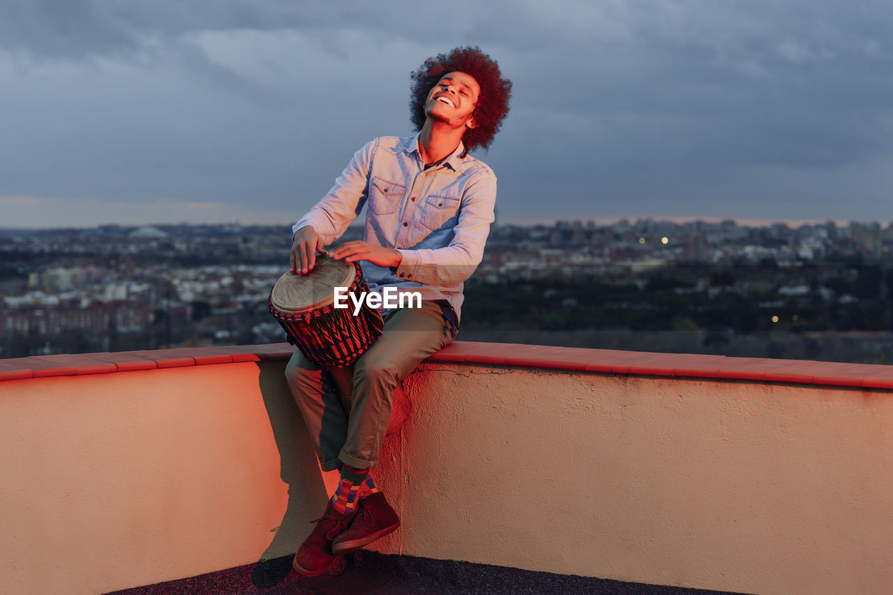 Young man singing and playing drum on terrace against cloudy sky at sunset