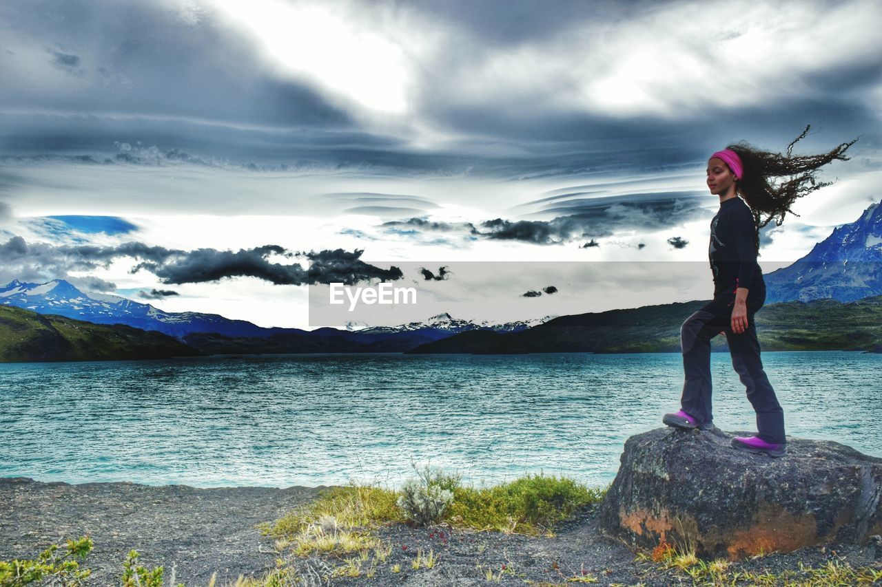 Girl standing on rock by sea against cloudy sky