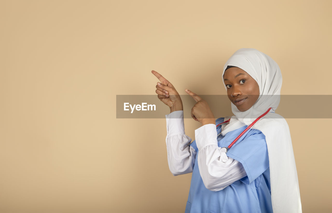 Smiling nurse in uniform and headscarf pointing fingers into empty space. advertising concept