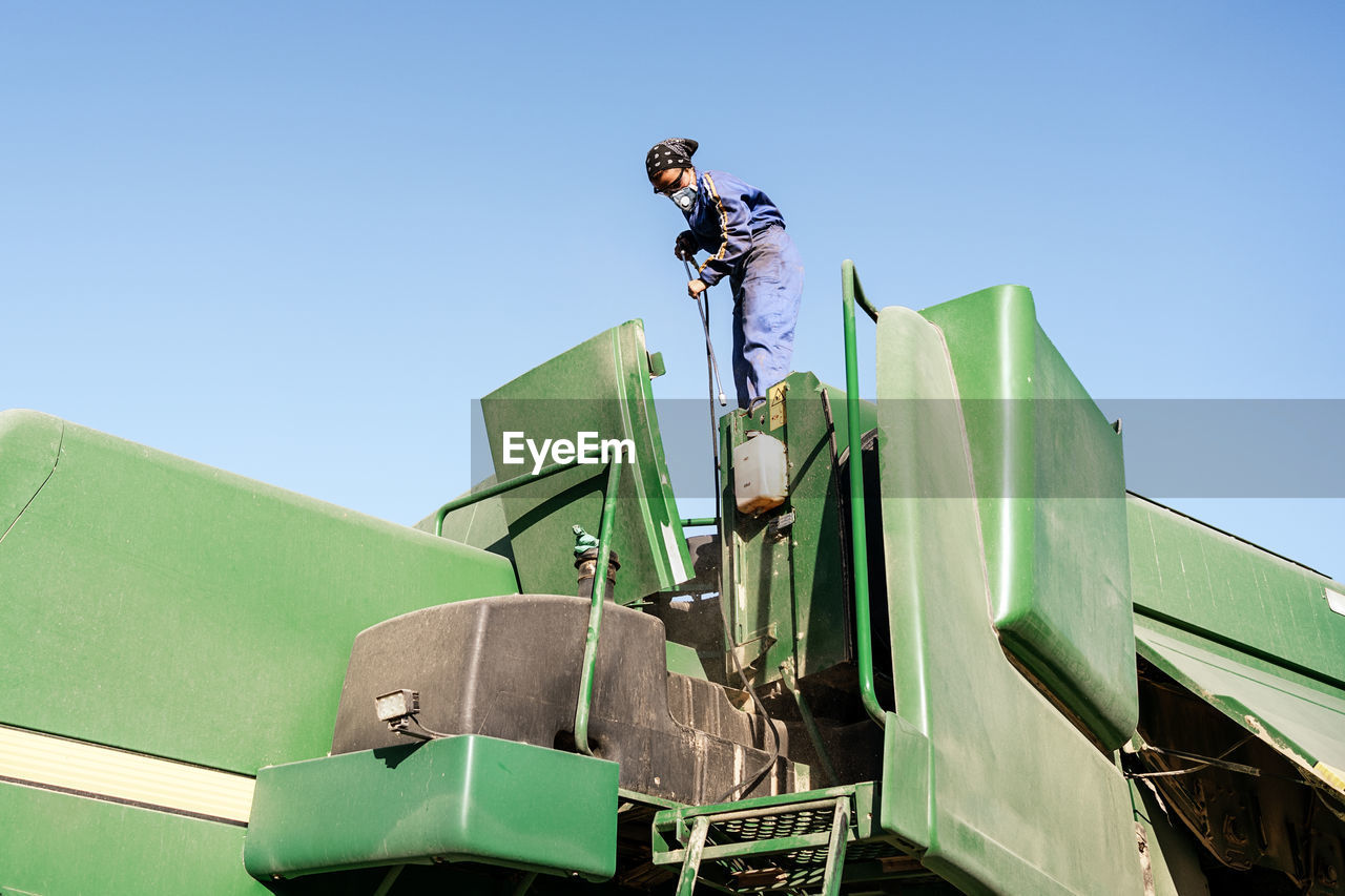 Low angle of unrecognizable employee in workwear and protective respirator cleaning agricultural machine in countryside on sunny day