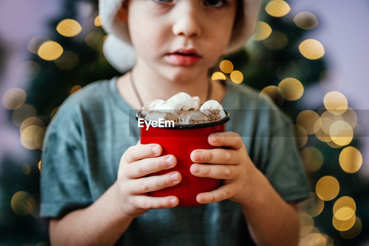 Little boy in santa hat holding a cup of hot chocolate with marshmallow