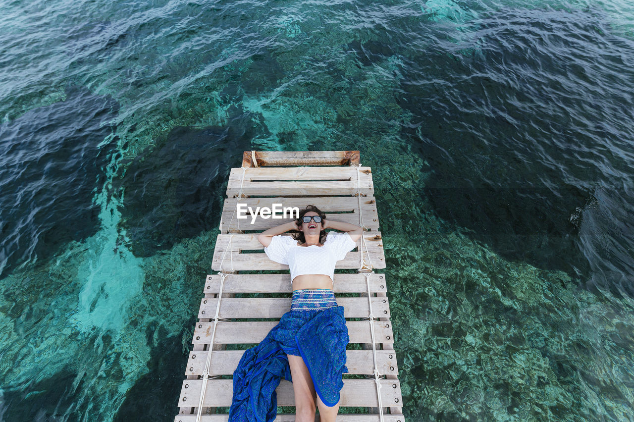 Happy woman with hands behind head lying on jetty during vacation