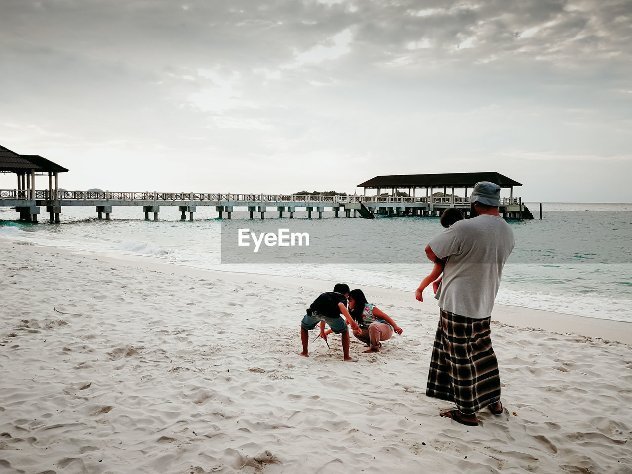 Father carrying daughter while looking at children playing on sand against sky