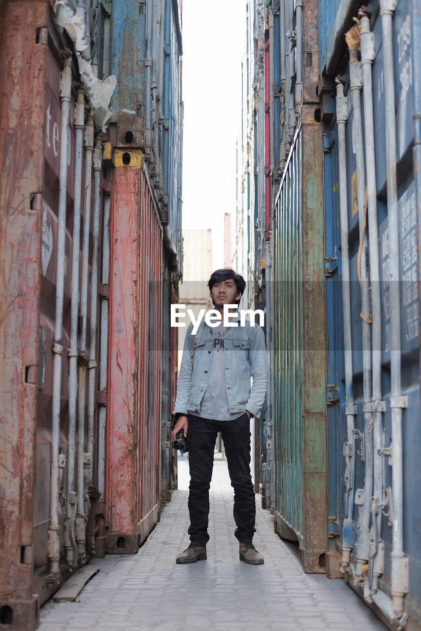 Full length of man holding camera while standing amidst cargo containers