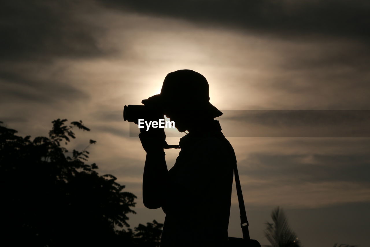 Side view of silhouette young man photographing against sky during sunset