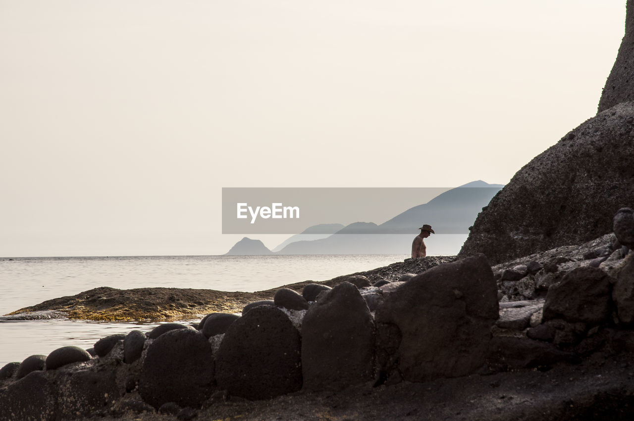 Man standing by rock formation at sea shore against clear sky