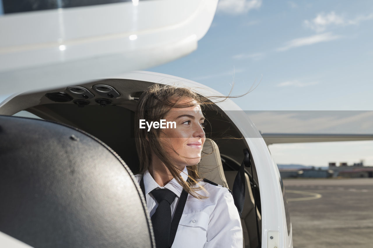 Smiling thoughtful female pilot looking away while sitting in airplane against sky at airport