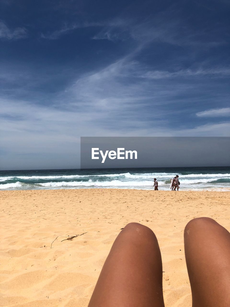 Cropped legs of woman relaxing at beach against blue sky