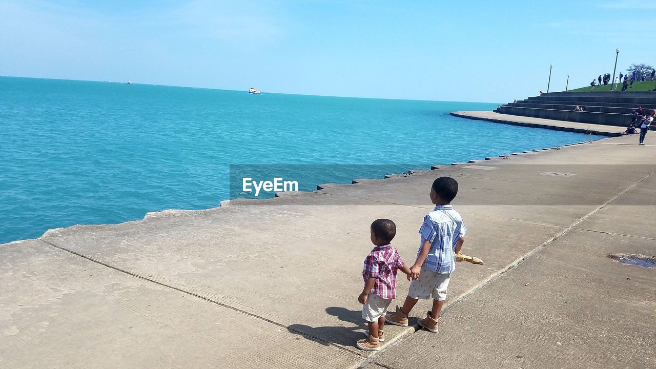 REAR VIEW OF BOYS PLAYING ON SEA AGAINST CLEAR SKY