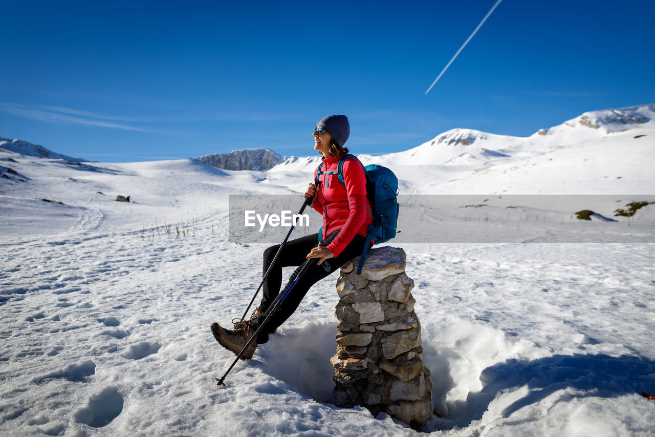 Woman rests and sunbathes sitting on a low wall, while hiking in the snow in the mountains. 