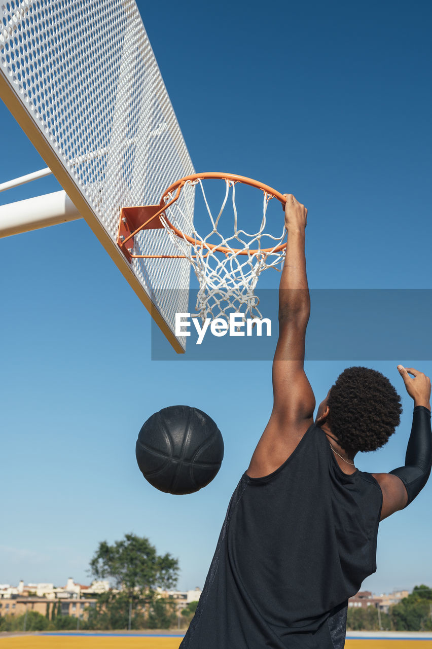 Back view of powerful energetic african american sportsman hanging on basketball lap after scoring ball in net on playground