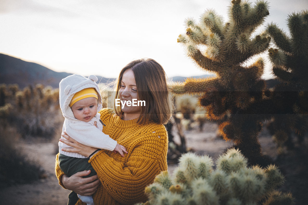 A woman with a baby is standing near a cactus in the desert