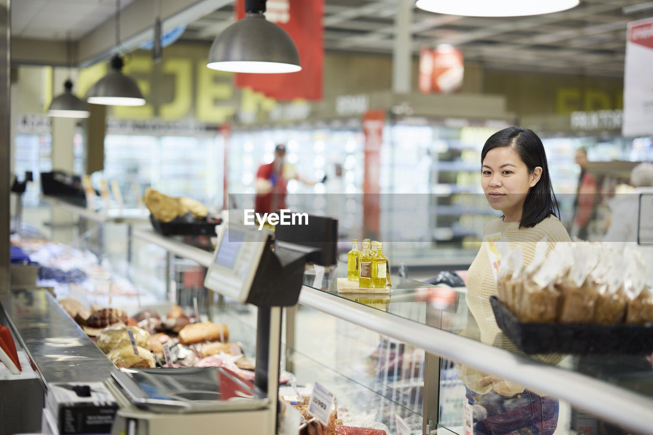 Female customer standing in front of deli counter in supermarket and checking products