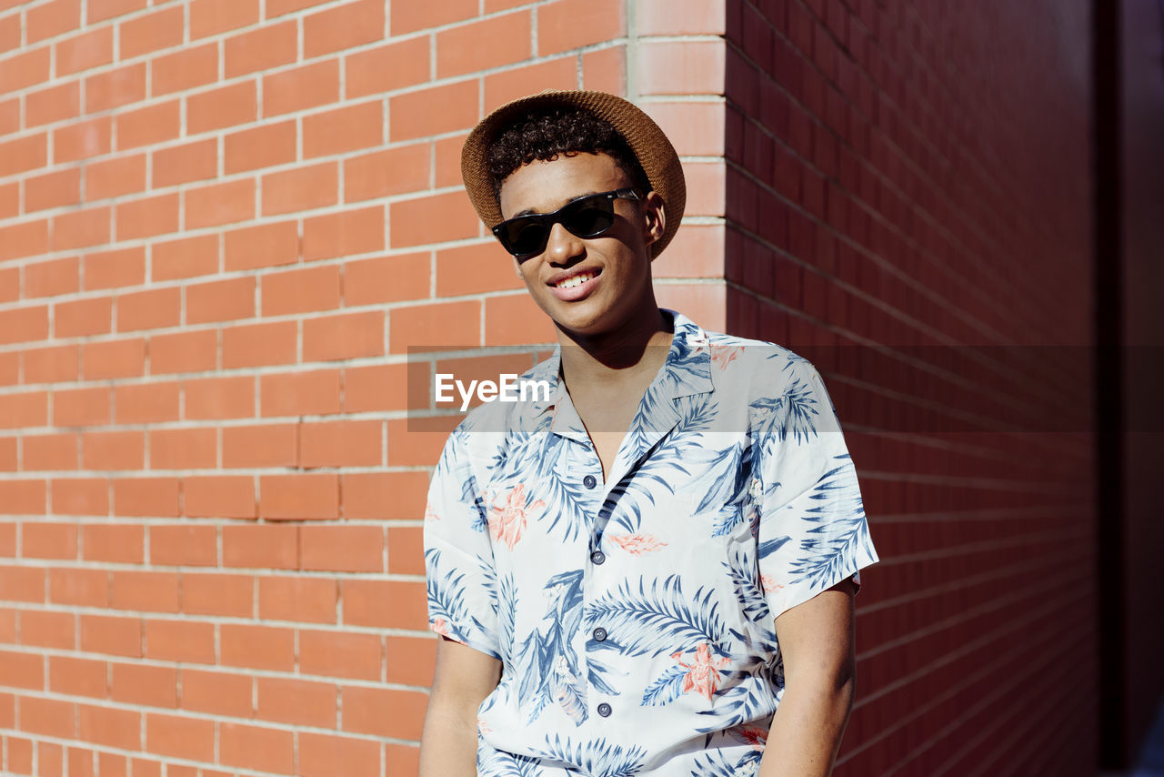 Trendy young black man with sunglasses and a hat posing on the street next to a brick wall looking at camera