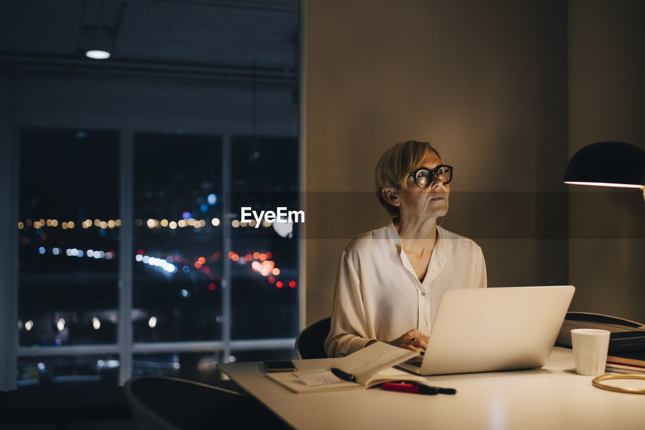 Thoughtful businesswoman sitting with laptop at illuminated desk in creative office