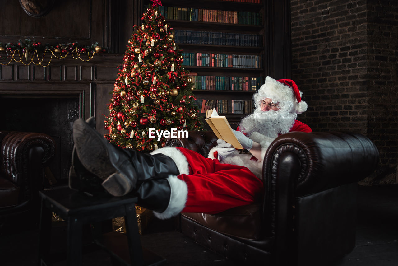Full length of man wearing santa claus costume reading book on sofa at home