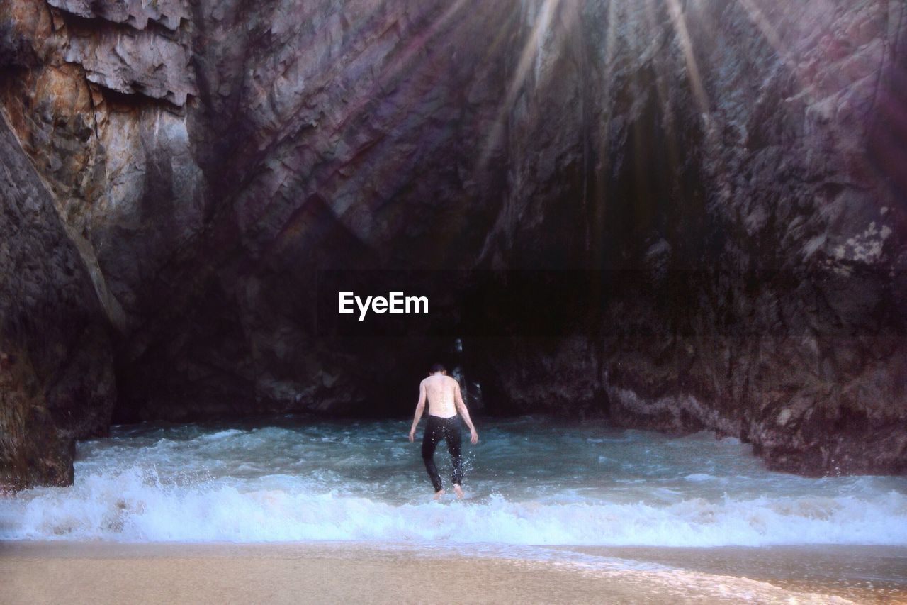 Rear view of shirtless man enjoying on shore against rock formations