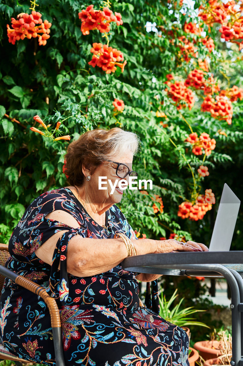 Concentrated elderly lady in casual outfit and eyeglasses sitting in chair at table while surfing on netbook near cup on saucer and green plants with flowers in yard in daylight